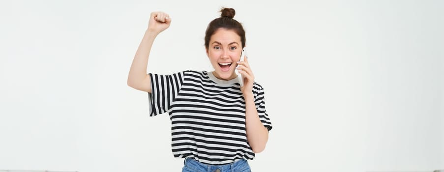 Portrait of enthusiastic young woman, talks on mobile phone, answers phone call and chanting, celebrating, laughing and smiling with excited face expression, white background.