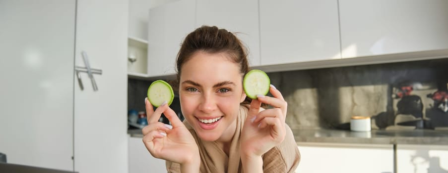 Portrait of beautiful brunette girl cooking in the kitchen, posing in bathrobe at home, holding zucchini, showing happy smile, making healthy food, vegetarian meal.