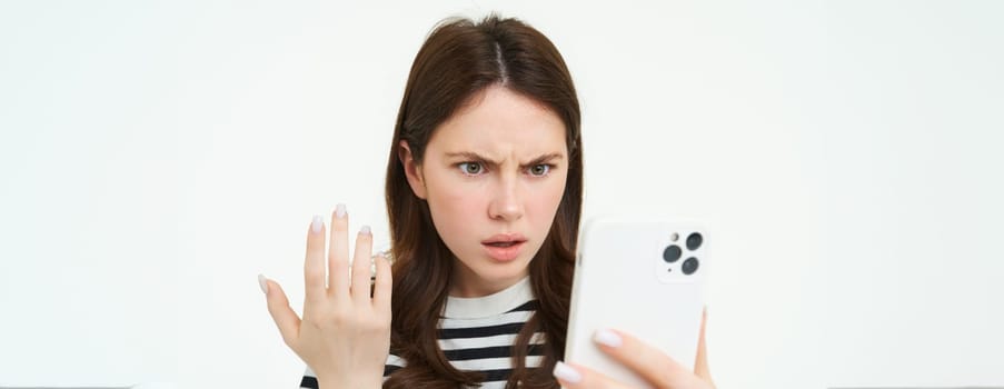 Portrait of annoyed girl staring at her mobile phone screen with frustrated face, white background.