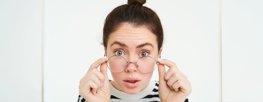 Portrait of woman leaning towards camera as if reading text with surprised face, takes off glasses, stands over white background. Copy space