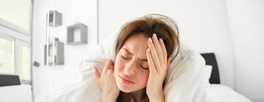 Portrait of woman with headache, lying in bed and touching head with frustrated, frowning face, does not feel well, stays in bedroom as having migraine in morning.