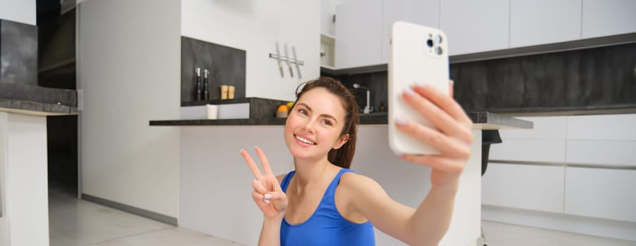 Close up portrait of beautiful brunette sportswoman, taking selfie on smartphone, posing on fitness mat with mobile phone, records her fitness exercises, workout training.