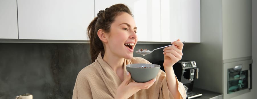 Gorgeous woman starts her day with cereals, eats breakfast and smiles, wears bathrobe, leans on kitchen worktop, starts her morning with quick meal.