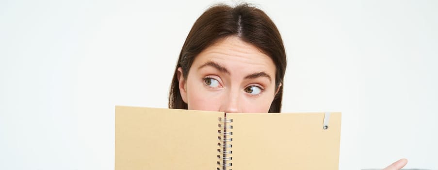 Close up portrait of woman with thoughtful expression, looking aside, hiding half of face behind notebook, standing with memo planner over white background.