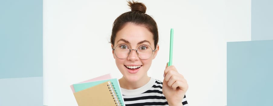 Portrait of woman in glasses, tutor raises hand with pen, eureka gesture, has idea or solution, holds notebook documents, stands over white background.