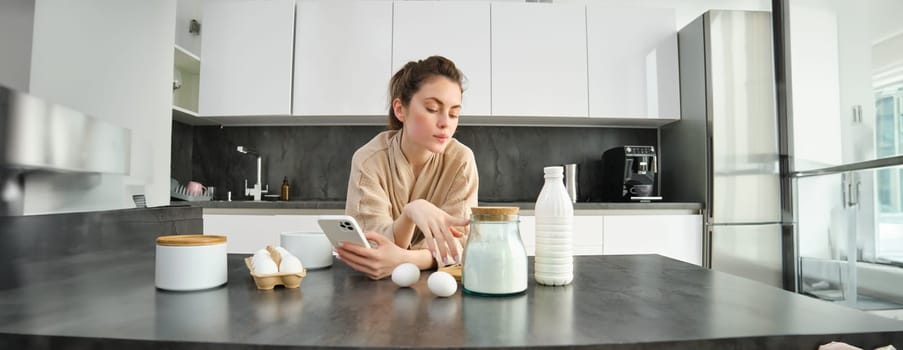 Attractive young cheerful girl baking at the kitchen, making dough, holding recipe book, having ideas.