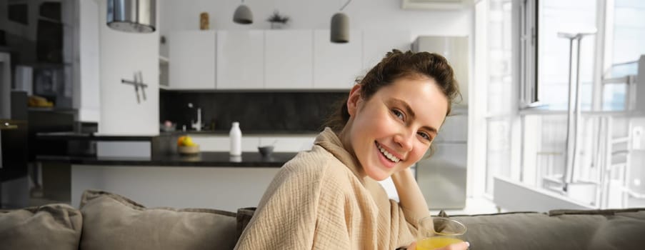 Women wellbeing and lifestyle. Beautiful young woman on sofa, drinking orange juice, relaxing at home on sofa, resting in the morning in living room.