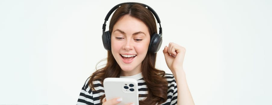 Cheerful, beautiful young woman using smartphone app, celebrating, looking happy while playing with mobile phone, listening music in wireless headphones.