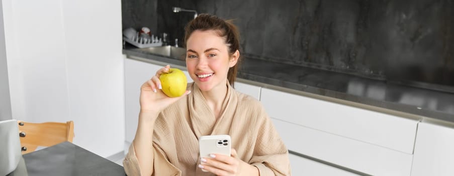 Close up portrait of smiling brunette woman in bathrobe, sits in kitchen at home, uses mobile phone and holds an apple, orders fresh fruits on smartphone app, buys groceries online, looks up recipe.