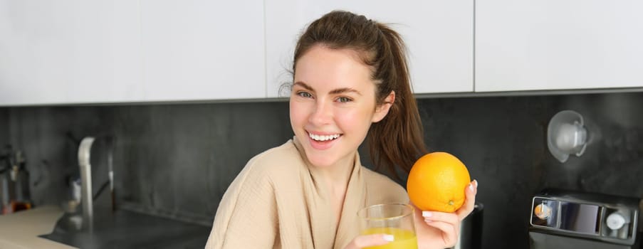 Close up portrait of stylish modern woman, drinking fresh juice from glass in kitchen, holding an orange, laughing and smiling, standing at home in bathrobe.
