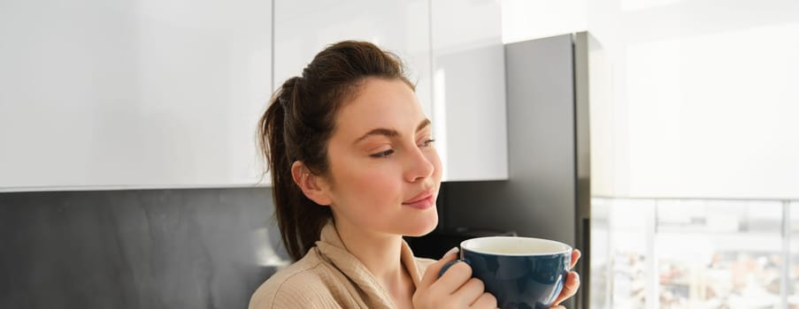 Daily routine and lifestyle. Young beautiful woman in bathrobe, standing in kitchen with cup of coffee, drinking tea, smiling and looking happy.