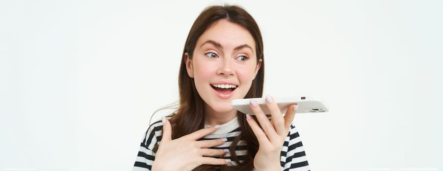 Image of cute brunette woman talking into speakerphone, holding mobile phone near mouth, records her voice, sends a voicemessage, using online translator app, white background.