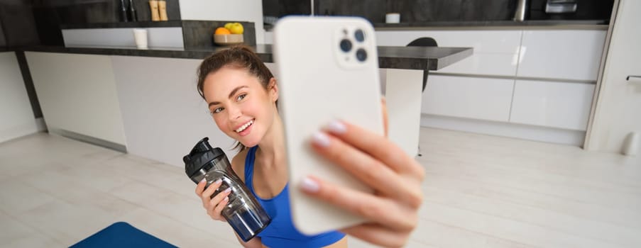 Portrait of sportswoman takes selfie with water bottle in living room, holds smartphone and poses for photo while doing fitness workout.