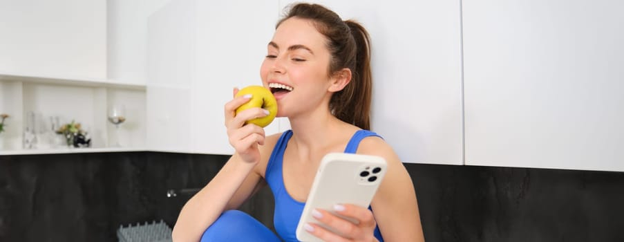 Image of stylish sportswoman, young fitness instructor sitting in kitchen and eating an apple, holding smartphone, using social media app on mobile phone.