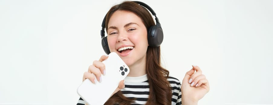 Technology and social media. Portrait of happy young woman singing karaoke, playing music app on smartphone, wearing wireless headphones, standing over white background.