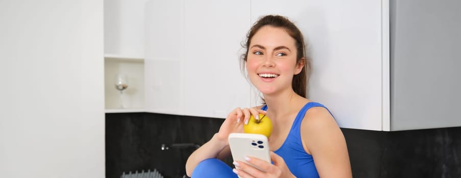 Portrait of stylish, young fitness woman, eating an apple and using mobile phone, holding smartphone, wearing sportswear.