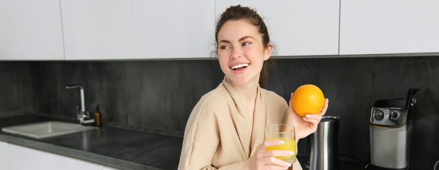 Portrait of good-looking brunette woman, drinking fresh orange juice in kitchen, holding fruit in hand, laughing and smiling, looking happy.