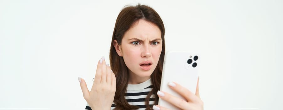 Portrait of confused, angry woman frowning, reading text on smartphone, watching video on mobile phone with shocked, puzzled face expression, isolated on white background.