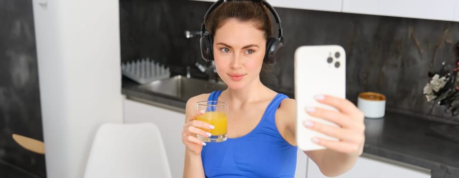 Portrait of young lifestyle, fitness blogger, woman with orange juice, takes selfie on smartphone camera, posing in activewear and wireless headphones.