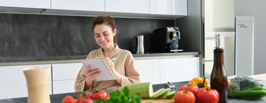 Portrait of happy young woman writes down menu for dinner, sits in the kitchen near vegetables, makes grocery list for shopping, poses in the bathrobe.