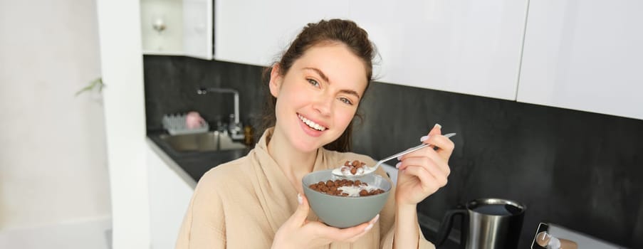 Close up of beautiful smiling woman in bathrobe, standing in kitchen near worktop, eating bowl of chocolate cereals with milk, holding spoon and looking happy at camera.
