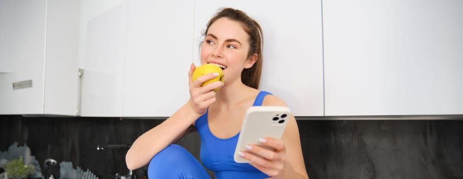 Portrait of healthy, sporty young woman, sitting in kitchen with smartphone, eating an apple, biting fruit.