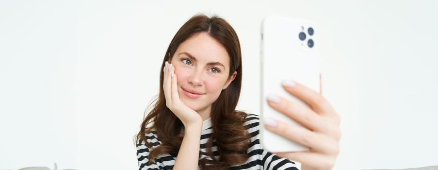 Portrait of young european woman taking selfie on smartphone, holding white mobile phone and posing for photos, isolated against white background.