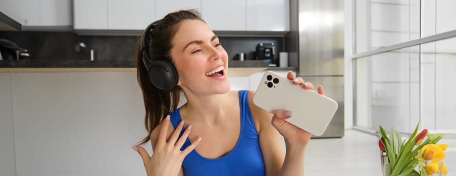 Close up portrait of happy, beautiful fitness woman, doing training exercises at home, wearing wireless headphones and singing at smartphone.