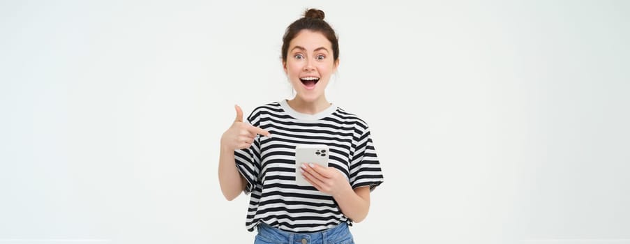 Portrait of beautiful young woman, recommending app on the phone, pointing at mobile screen, showing smth on smartphone, standing against white background.