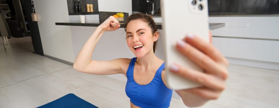 Portrait of active fitness girl, takes selfie on smartphone during home workout, posing on fitness mat, shows her strong muscles and laughs.