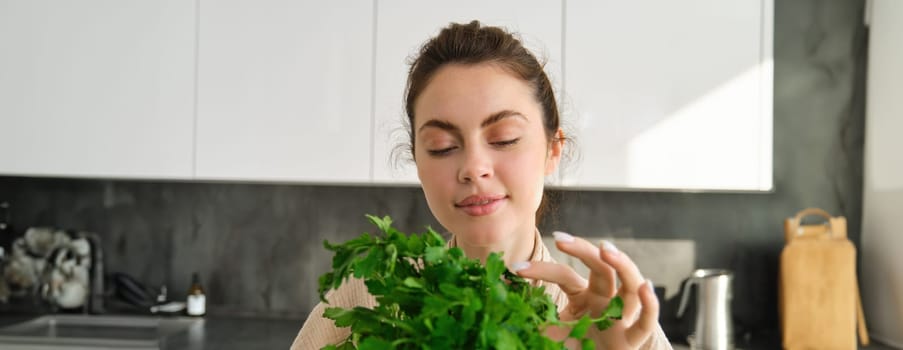 Portrait of attractive girl with bunch of parsley, eating fresh herbs and vegetables, cooking in the kitchen.