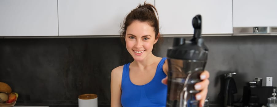 Portrait of beautiful young fitness girl, offering you water bottle to drink after workout, standing in kitchen, smiling at camera.