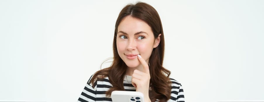 Portrait of smiling woman thinks to order something online on smartphone app, holding mobile phone with thoughtful face.