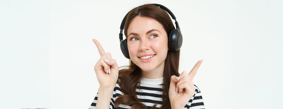 Close up portrait of beautiful girl dancing, listening music in wireless headphones and smiling, pointing sideways, showing two sides, advertisement or banner.