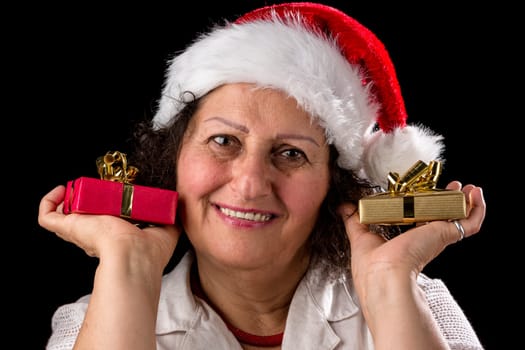 Venerable pensioner with a Santa hat smiles. She holds a small Christmas parcel in each hand,one golden,the other wrapped in red. Black background. Wide depth of field. Venerable middle-aged woman with a Kris Kringle has cracking a smile. She is holding a small Xmas present in each hand,wrapped in gold and red. Black background. Focus on eyes. Ample depth of field.
