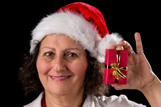 Smiling mature woman with a red Father Christmas cap is holding up her left hand and offering a small wrapped present. Pitch black background. Focus on the eyes. Medium depth of field. Teeth show. Smiling senior woman with a red Christmas hat holds and shows a small gift packet with her left hand. Black background. Focus on the eyes. Medium depth of field. Teeth are visible.