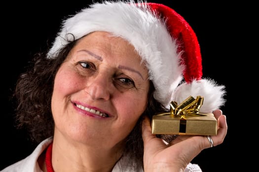 Smiling mature woman with a Santa Claus cap holding a Christmas present all wrapped in gold next to her left cheek. Pitch black background. Ample depth of field with focus on her eyes. Smiling mature lady with a Christmas hat holds a gold-packed Christmas gift near her left cheek. Pitch black background. Wide depth of field with focus on the eyes.