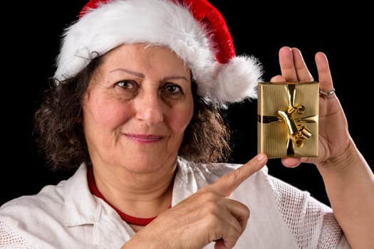 Friendly,venerable,dark-haired woman wearing an Old Father Christmas cap. She is pointing with her right index finger at a small golden Xmas present that she is holding up with her left hand. Friendly,venerable,dark-haired woman wears a Santa hat. She points her right index finger at a golden gift,which she holds up with her left hand.