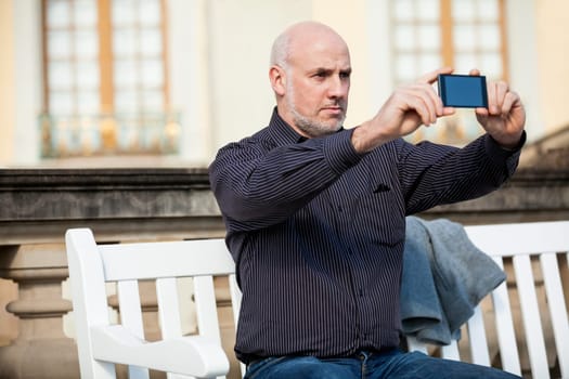 Attractive older man senior photographs himself with his mobile phone smartphone in the park