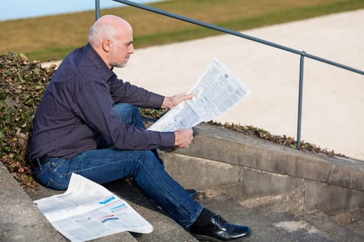 Middle-aged man,senior sitting with a newspaper on a staircase in spring outdoors