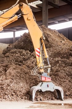 Excavator with gripper arm in front of a large pile of brown earth in the construction industry
