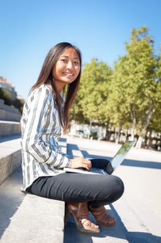 Happy asian girl using a laptop sitting on stairs in a park