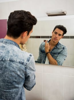 Attractive young man looking at bathroom mirror, spraying cologne or perfume