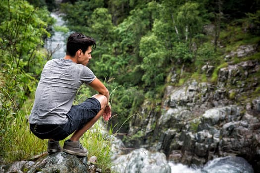 Handsome muscular young man sitting in lush green mountain scenery looking down at water stream