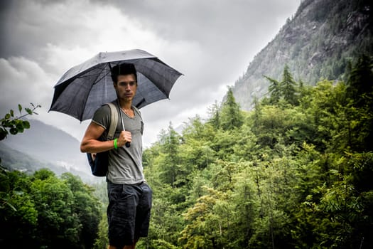 Smiling handsome young man holding an umbrella on a stormy day as he takes a walk along a trail in the mountains, looking at camera