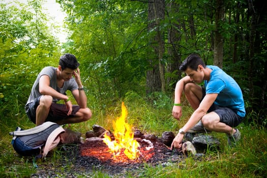 Two young men camping in the mountains crouching alongside a burning campfire with rucksack staring thoughtfully at the fire