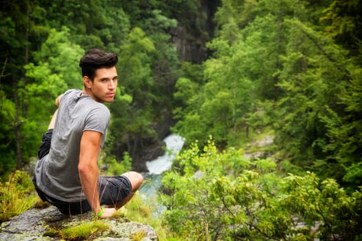 Handsome muscular young man sitting in lush green mountain scenery looking at camera
