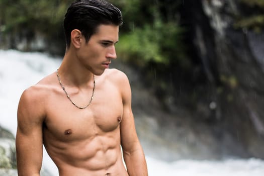 Handsome shirtless young man near mountain waterfall, with ripped fit body