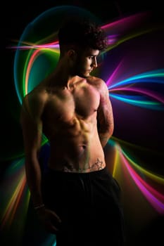 Young athletic shirtless man with tattoo posing over dark colorful background, studio shot