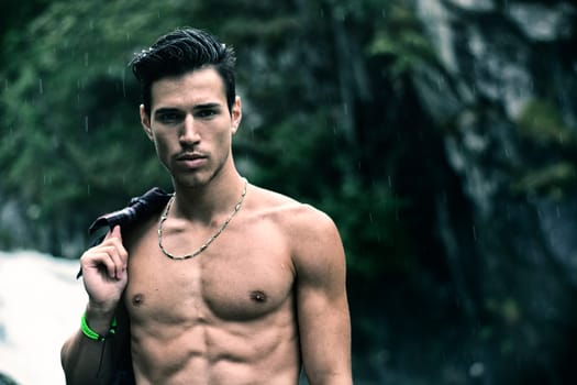 Handsome young man near mountain waterfall shirtless, under rain